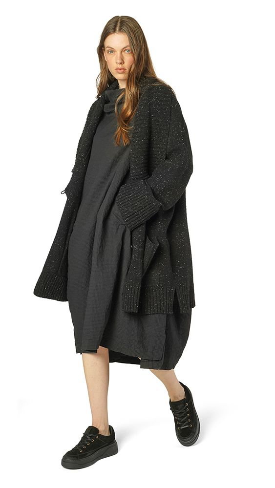 Frau trägt oversized schwarzen Strickmantel mit Knopfdetails, kurze schwarze Hose und Sneakers.
