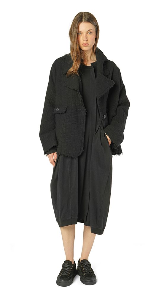 Schwarzer oversized Mantel mit Fransen über schwarzem Kleid, kombiniert mit schwarzen Sneakern.