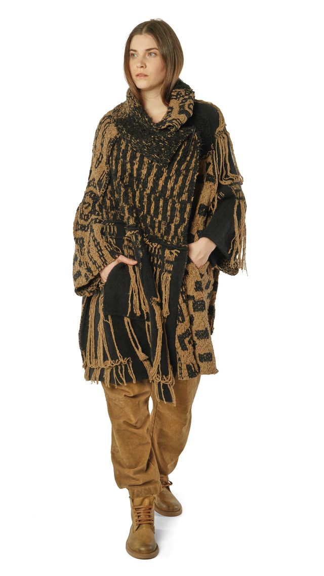 Frau trägt oversized gestrickten Poncho mit Fransen in Beige-Schwarz, dazu beige Hosen und Stiefel.
