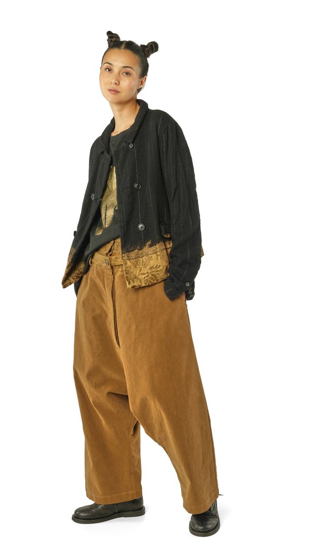 Person trägt oversized schwarze Jacke, goldbraune Hosen und Accessoires mit bunten Stickereien, Haare zu zwei Buns gebunden.