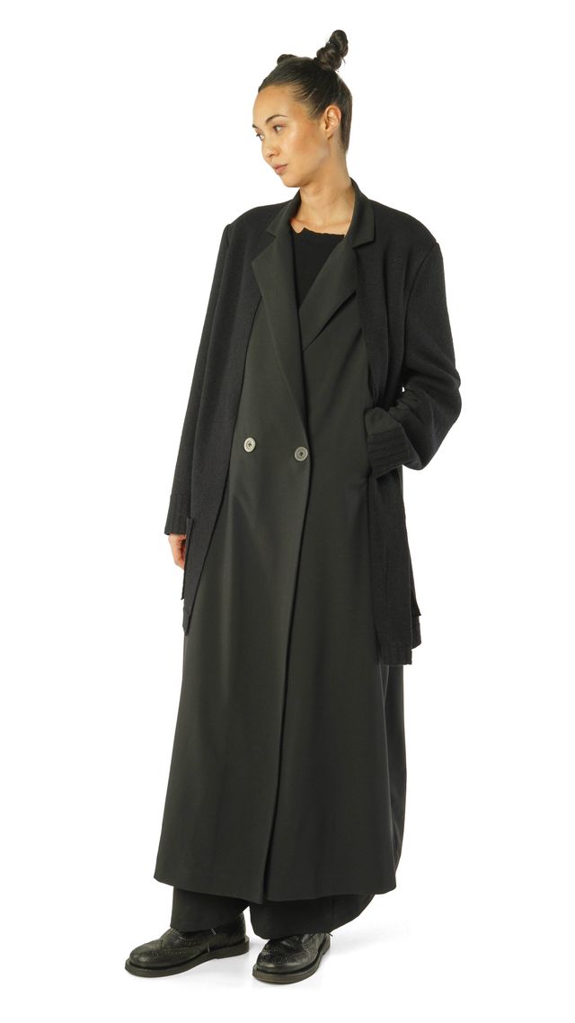 Schlanke Person in langem schwarzem Trenchcoat mit schwarzen Hosen und Stiefeln.