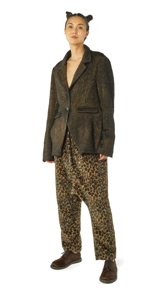 Person trägt oversized Tweedjacke, leopardgedruckte Hosen und Kopfband mit Ohren, barfuß auf hellen Untergrund.