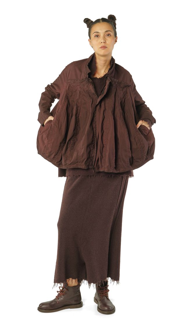 Person trägt oversized braunen Mantel mit weitem Rock und robusten Stiefeln, Haare zu zwei Hörnern gebunden.
